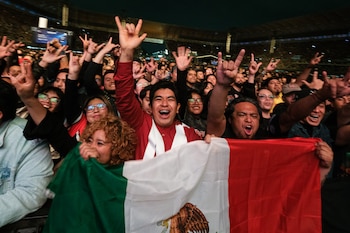 Personas en la multitud de un estadio sostienen una bandera de México en primer plano y hacen señas de rock con los dedos levantados