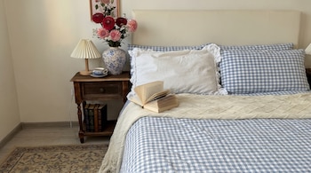 Una cama con ropa de cama a cuadros azul y blanco, almohadas, manta, libro abierto, mesita de noche de madera, lámpara, taza, jarrón con flores y libros.