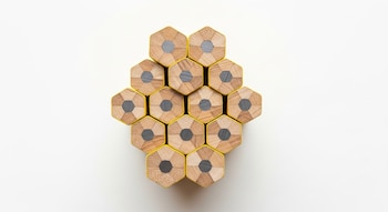 Vista cenital de doce lápices hexagonales de madera y amarillo apilados en un patrón de colmena sobre un fondo blanco, mostrando los extremos de grafito.