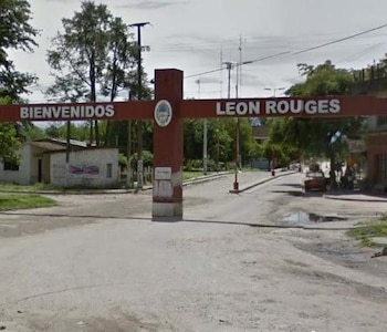 Estructura de acceso roja con el cartel Bienvenidos León Rouges sobre una calle asfaltada rodeada de árboles y edificaciones