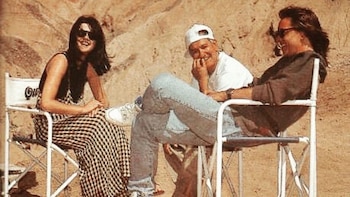 Tres personas, una mujer y dos hombres, sentados en sillas de director blancas. La mujer lleva gafas de sol, un hombre una gorra blanca. Fondo arenoso