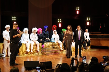 Moria Casán, en un vestido marrón, de pie en un escenario. Detrás, artistas con pelucas de colores sentados en un podio dorado. Un hombre con micrófono, público filma