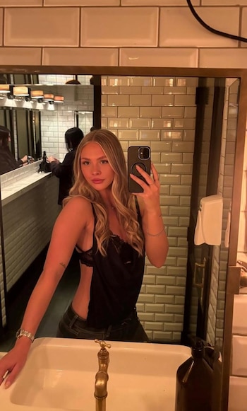 Mujer rubia toma un autorretrato en un espejo con un teléfono móvil. En el fondo hay azulejos en la pared, un lavabo y un grifo. Se ve una persona reflejada