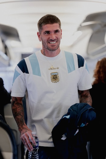 Hombre joven sonriente, pelo corto y barba, camiseta blanca con franjas celestes y azules, escudo AFA, collar, tatuajes en brazos, botella de agua, interior de avión