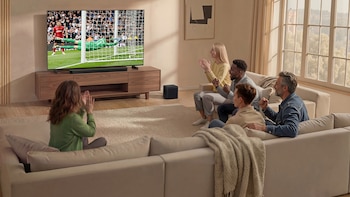 Cinco personas en una sala de estar, sentadas en sofás beige, viendo un partido de fútbol en un televisor grande Samsung con una barra de sonido. Un portero ataja un gol