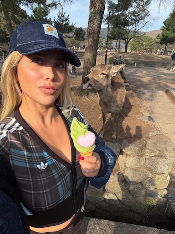 Wanda Nara, rubia y con gorra azul, se toma una selfie sonriendo mientras sostiene un helado verde y rosa. Detrás, un ciervo saca la lengua