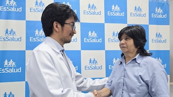 Un doctor sonriente con bata blanca y gafas le da la mano a una mujer con camisa azul claro, ambos frente a un fondo azul y blanco con logos de EsSalud