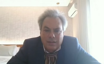Primer plano de un hombre con cabello gris, traje azul, camisa blanca y corbata a rayas. Fondo con pared clara estampada, cama y aire acondicionado