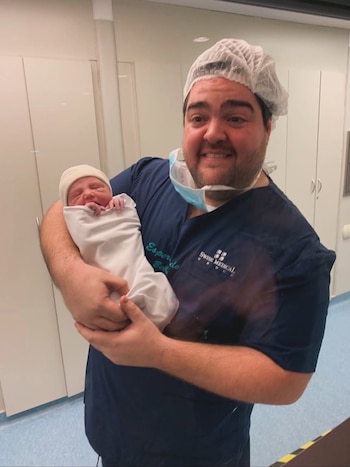 Darío Barassi sonriente, vestido con uniforme médico azul y gorro, sostiene a un bebé recién nacido envuelto en blanco y con gorro claro