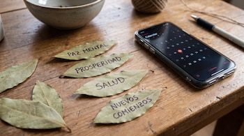Hojas de laurel con texto escrito sobre mesa de madera. Un teléfono celular con calendario digital, un bolígrafo, un cuenco y un ovillo de hilo.