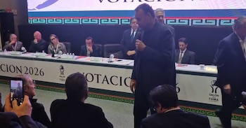 David Faitelson, vestido de negro, de pie y hablando por un micrófono en un evento. Detrás, una mesa con personas y un letrero de "Salón de la Fama"