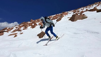 Aron Ralston esquía por una ladera montañosa cubierta de nieve y rocas marrones, vestido con chaqueta gris, pantalones azules y mochila, bajo un cielo azul claro