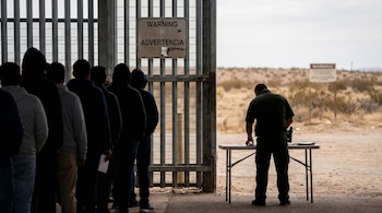 Personas en fila frente a una valla. Un agente revisa documentos en una mesa. Se observan letreros de advertencia y un paisaje desértico.