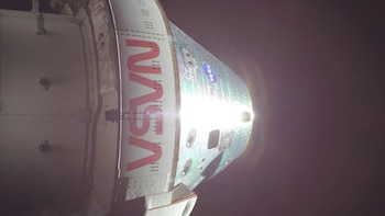 Vista de cerca de la cápsula Orion de la NASA en el espacio, con el logotipo rojo de NASA visible y una luz brillante reflejándose en su superficie