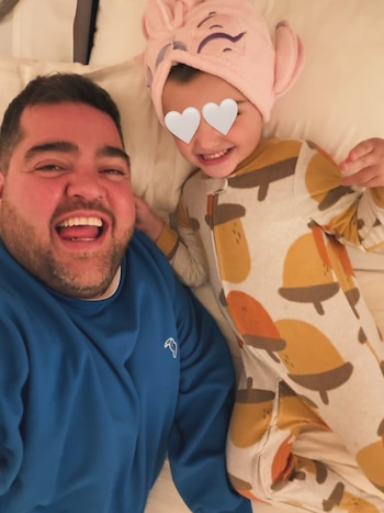 Un hombre con barba y sonrisa, vestido con un buzo azul, recostado junto a una niña con pijama estampado, gorro de toalla y ojos cubiertos con corazones