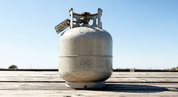 Un tanque de gas LP de tamaño pequeño y color claro, con una etiqueta que muestra '2026' y los números '03' y '09', descansa sobre tablas de madera bajo un cielo azul claro.