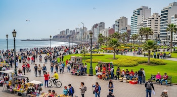 Gran cantidad de personas caminando y comprando en el malecón de Miraflores, Lima. Se ven puestos de venta, palmeras, edificios y el mar con parapentes.