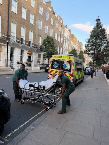 Dos paramédicos empujan una camilla con una persona envuelta en una sábana blanca cerca de una ambulancia amarilla y verde estacionada en una calle urbana