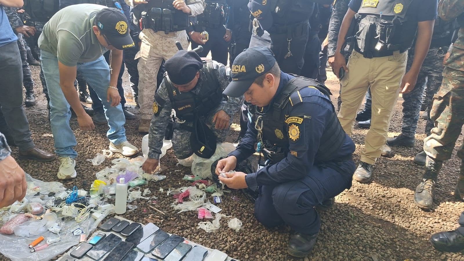 Agentes policiales y personal civil examinan una colección de teléfonos móviles, encendedores y envoltorios plásticos sobre el suelo de grava durante un operativo, en la Cárcel Renovación I (PNCdeGuatemala)