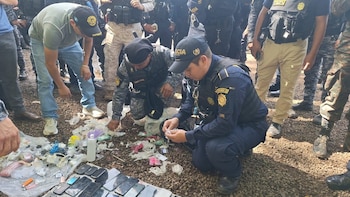Hombres con uniformes y civiles examinan teléfonos móviles, encendedores, cables, una botella y bolsas plásticas sobre un suelo de grava