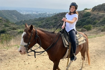 Una mujer con casco monta un caballo marrón y blanco por un sendero de tierra, con colinas y una zona urbana en el fondo