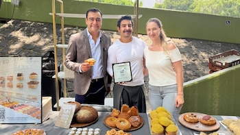 Mauricio Tabe posa entre un panadero y una mujer, con una mesa llena de diversos panes y productos de repostería artesanal en un evento al aire libre