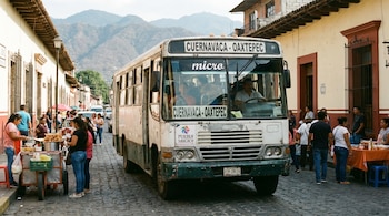 Un autobús blanco y verde transita por una calle empedrada con personas. Muestra letreros que dicen CUERNAVACA - OAXTEPEC, micro y Pueblo Mágico.