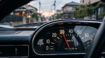 Tablero de automóvil con velocímetro analógico que marca 30 kilómetros por hora y una calle residencial desenfocada al fondo.