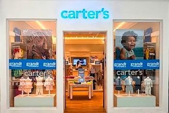 Fachada de una tienda Carter's con su letrero azul brillante, dos grandes escaparates con ropa de bebé y un banner azul que dice 'grand opening'