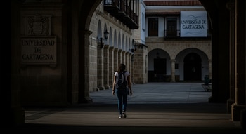 Una estudiante con mochila camina de espaldas a través de un arco oscuro hacia un edificio con arcos y un cartel de "Universidad de Cartagena".