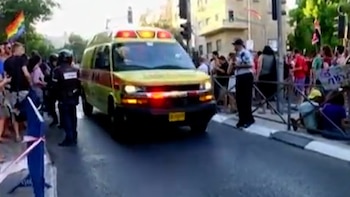 Una ambulancia con luces encendidas en una calle con personas. Una bandera arcoíris se ve entre la multitud. Hay edificios y árboles al fondo