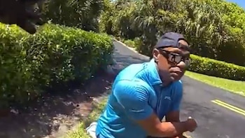 Un hombre con gorra y gafas de sol, vistiendo una camiseta azul, se inclina junto a una carretera con arbustos y árboles verdes de fondo