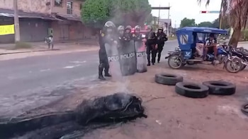 Oficiales de policía con escudos antidisturbios se paran en una calle con humo y objetos quemándose en primer plano; una mototaxi está estacionada cerca