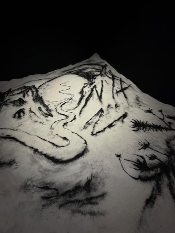Una maqueta o superficie blanca con arena negra esparcida formando un paisaje montañoso, un río sinuoso y elementos vegetales sobre un fondo negro