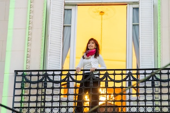 Cristina Kirchner, con cabello castaño rojizo y un pañuelo de colores, sonríe desde un balcón con barandal oscuro y un fondo amarillo brillante