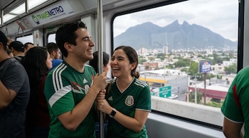 Dos personas sonrientes con camisetas verdes de la selección mexicana viajan en el Transmetro de Monterrey, con una ciudad y montañas visibles por la ventana.
