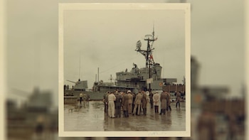 Grupo de personal militar en uniformes de pie sobre una cubierta mojada, con un gran buque de guerra gris, el USS Hornet, al fondo bajo un cielo nublado