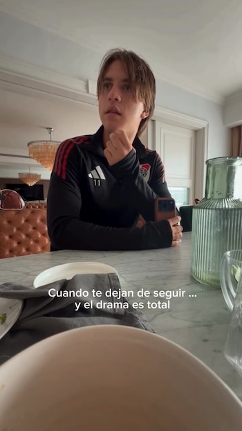 Un joven con camiseta negra de River Plate y cabello castaño mira pensativo hacia arriba, sentado en una mesa de mármol con un teléfono en la mano