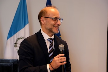 Un hombre sonriente con anteojos, traje y corbata sostiene un micrófono frente a una bandera de Guatemala