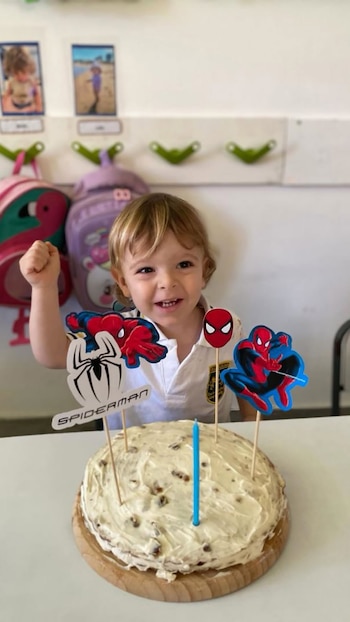 Un niño rubio sonriente con camiseta blanca posa junto a una torta blanca con trozos y decoraciones de Spiderman, incluyendo una vela azul