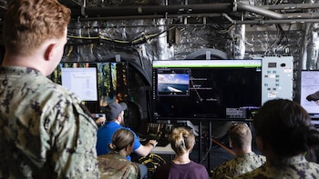 Varias personas, incluyendo personal militar, observan pantallas de computadora. Una pantalla grande muestra una embarcación en el mar con un mapa digital