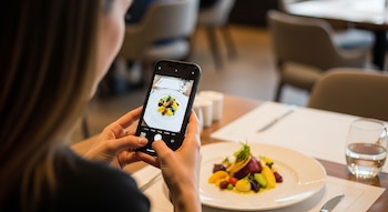 Mujer con teléfono móvil fotografiando un plato de comida en una mesa de restaurante. Hay cubiertos, un vaso de agua y mesas al fondo.