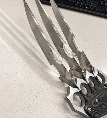 Un arma metálica de puño con tres hojas puntiagudas apoyada sobre una mesa blanca junto a un teclado negro de computadora