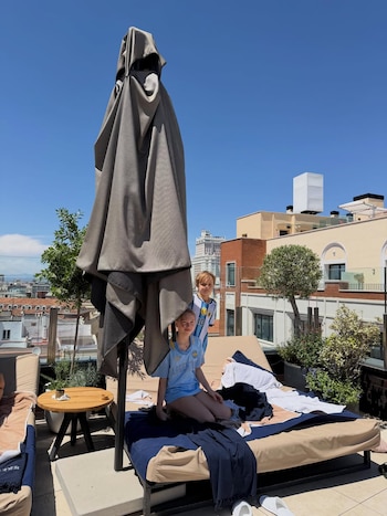 Dos niños con camisetas de Argentina en tumbonas sobre una terraza. Se observa un paraguas plegado, una mesa y edificios de ciudad bajo cielo azul despejado