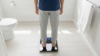 Un hombre de espaldas se para sobre una báscula digital que muestra 75.2 kg. En el baño se observan un inodoro, un rollo de papel y una toalla.