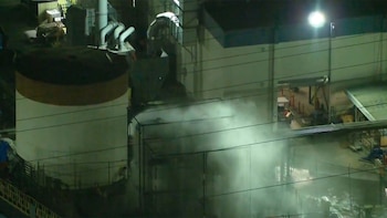 Vista nocturna de una instalación industrial con vapor blanco emergiendo de una estructura central y cubriendo una parte del área, bajo luces artificiales