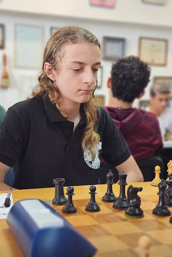 Luciano Curcio, un joven con cabello largo y camisa negra, sentado frente a un tablero de ajedrez con piezas y un reloj azul