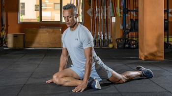 Un hombre de mediana edad con camiseta gris y pantalones cortos azules se estira en el suelo de un gimnasio con equipo deportivo al fondo.