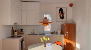 Vista de una cocina moderna con gabinetes superiores blancos, inferiores beige, una nevera naranja estilo retro, una lámpara colgante y una mesa redonda con flores.
