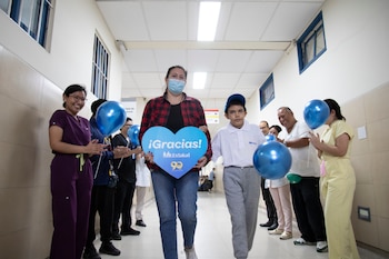 Un niño con gorra y una mujer que sostiene un cartel azul en forma de corazón caminan entre trabajadores de salud que aplauden en un pasillo